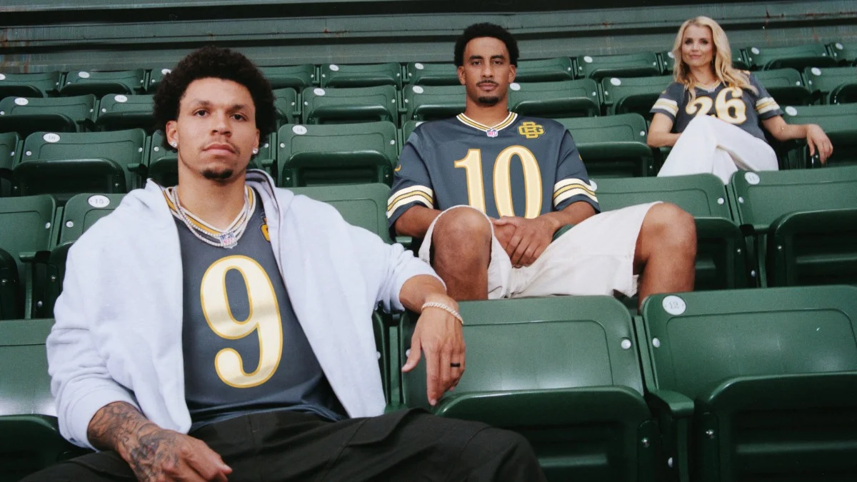 Drei Personen, die graue Nike-Sporttrikots mit den Nummern 9, 10 und 26 tragen, sitzen mit Abstand zueinander auf leeren grünen Stadionplätzen und präsentieren die „RivalriesKollektion“, die von legendären NFL-Duellen inspiriert ist. Alt-Text wurde KI-generiert.
