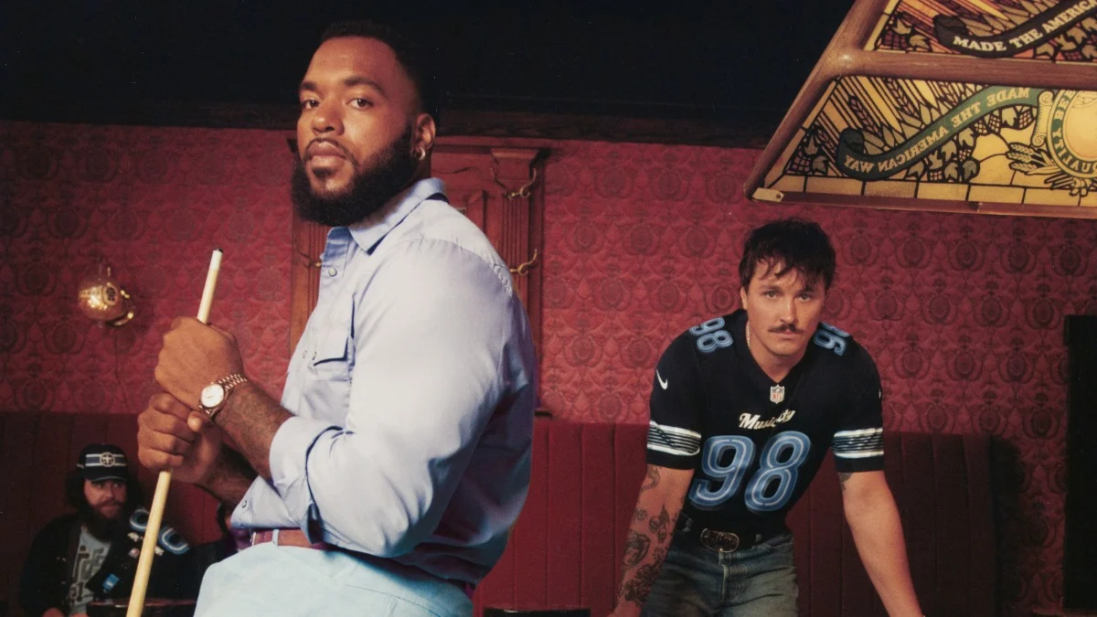 Zwei Männer stehen in einer schummrig beleuchteten Bar; einer hält einen Billardqueue in der Hand, während der andere, der ein NFL-Trikot aus der Nike Rivalries-Kollektion trägt, sich nach vorne beugt. Im Hintergrund sitzt eine dritte Person. Alt-Text wurde KI-generiert.