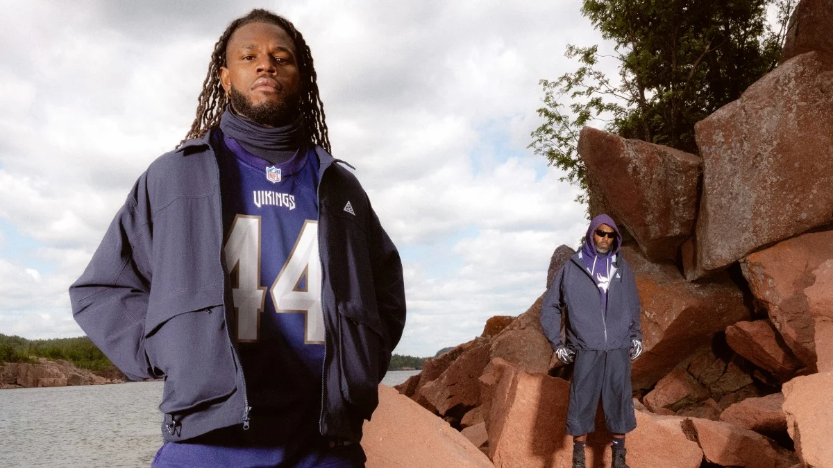 Zwei Personen stehen auf großen, rötlichen Felsen am Wasser; die Person im Vordergrund trägt ein NFL-Trikot und eine Jacke der Vikings, während die andere eine Kapuzenjacke und eine Sonnenbrille trägt. Alt-Text wurde KI-generiert.