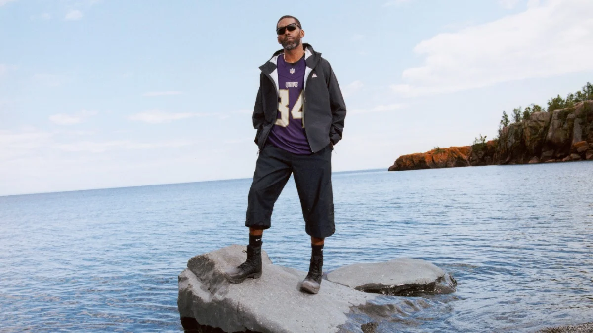 Eine Person, die eine Sonnenbrille, ein Nike-Sporttrikot, eine Jacke, eine kurze Hose und Stiefel trägt, steht auf einem Felsen, der von Wasser umgeben ist, im Hintergrund sind eine entfernte Küstenlinie und ein blauer Himmel zu sehen. Alt-Text wurde KI-generiert.