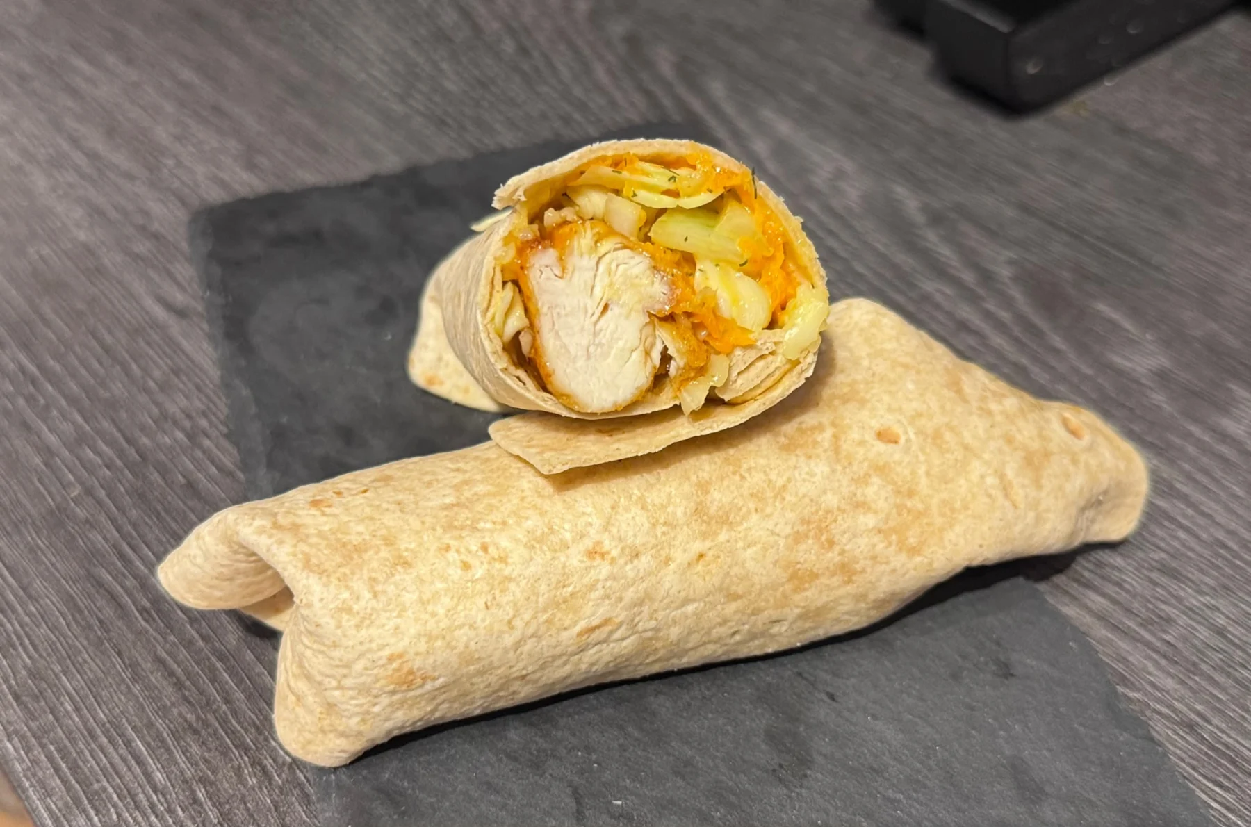 Ein halbierter „Buffalo Chicken Burrito“-Wrap zeigt eine Füllung aus Hähnchenfleisch, Kohl und Möhren, die auf einem dunklen Untergrund liegt. Alt-Text wurde KI-generiert.