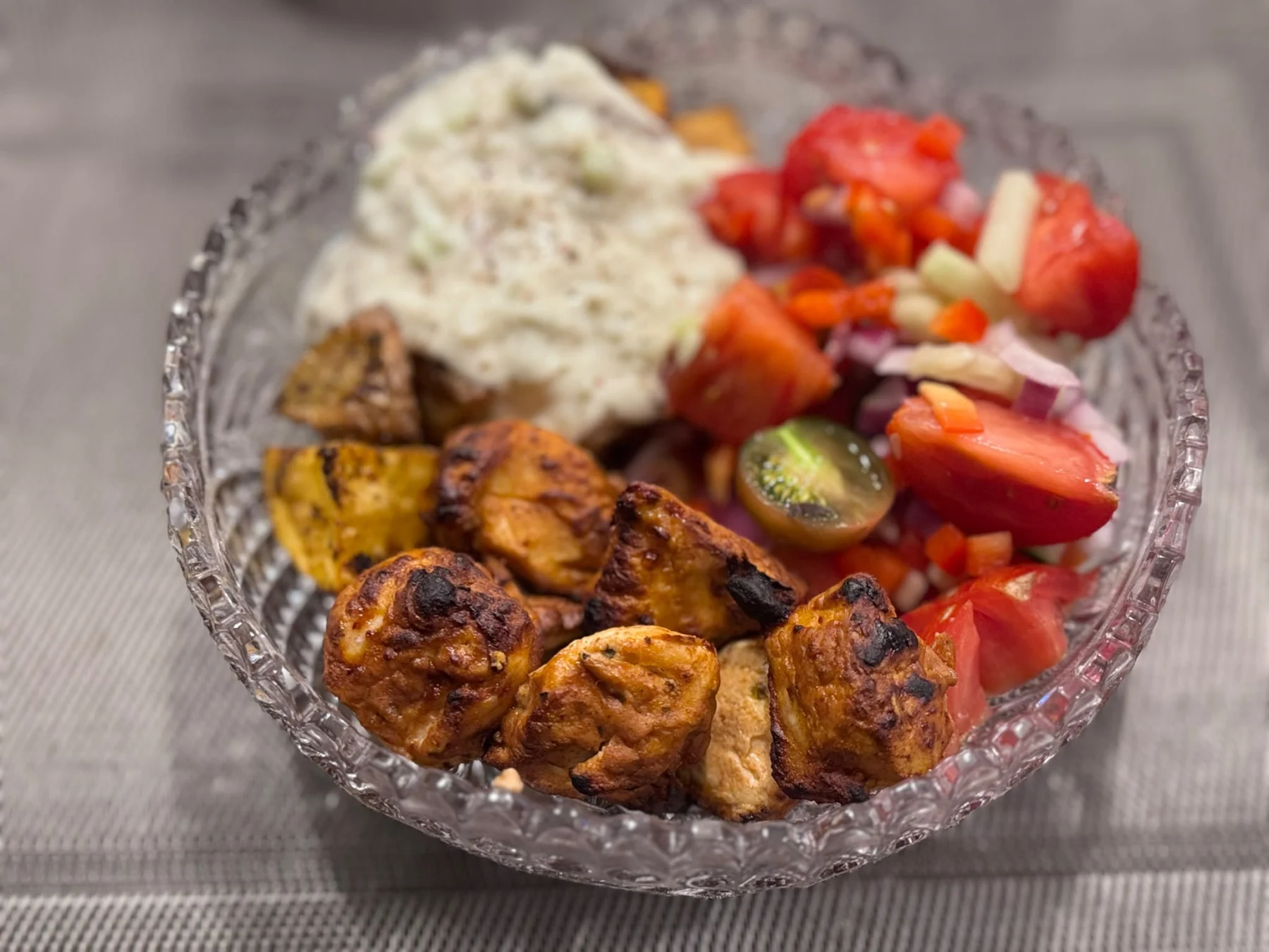 Eine Glasschüssel mit gegrillten Hähnchenstücken, Bratkartoffeln, einem Tomaten-Gurken-Salat und einer Portion Tzatziki – diese Chicken-Souvlaki-Bowl vereint klassische mediterrane Aromen zu einer frischen und sättigenden Mahlzeit. Alt-Text wurde KI-generiert.