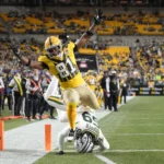 Ein Footballspieler in einem gelben Trikot, das an die Packers erinnert – die kürzlich Jonnu Smith verpflichtet haben –, überquert die Seitenlinie, während ein Verteidiger in einem weißen Trikot ihn nahe der Endzone während eines Spiels in einem Stadion tackelt. Alt-Text wurde KI-generiert.