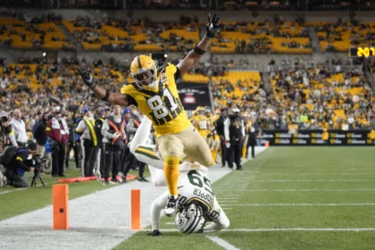 Ein Footballspieler in einem gelben Trikot, das an die Packers erinnert – die kürzlich Jonnu Smith verpflichtet haben –, überquert die Seitenlinie, während ein Verteidiger in einem weißen Trikot ihn nahe der Endzone während eines Spiels in einem Stadion tackelt. Alt-Text wurde KI-generiert.