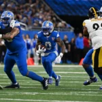 Jahmyr Gibbs, ein Runningback der Detroit Lions, läuft mit dem Football, während seine Teamkollegen und die Verteidiger der Pittsburgh Steelers während eines NFL-Spiels in einen Zweikampf verwickelt sind. Alt-Text wurde KI-generiert.
