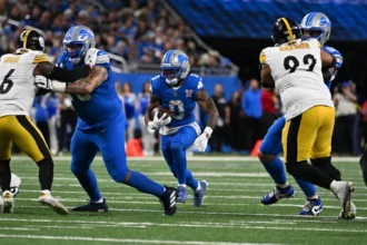 Jahmyr Gibbs, ein Runningback der Detroit Lions, läuft mit dem Football, während seine Teamkollegen und die Verteidiger der Pittsburgh Steelers während eines NFL-Spiels in einen Zweikampf verwickelt sind. Alt-Text wurde KI-generiert.