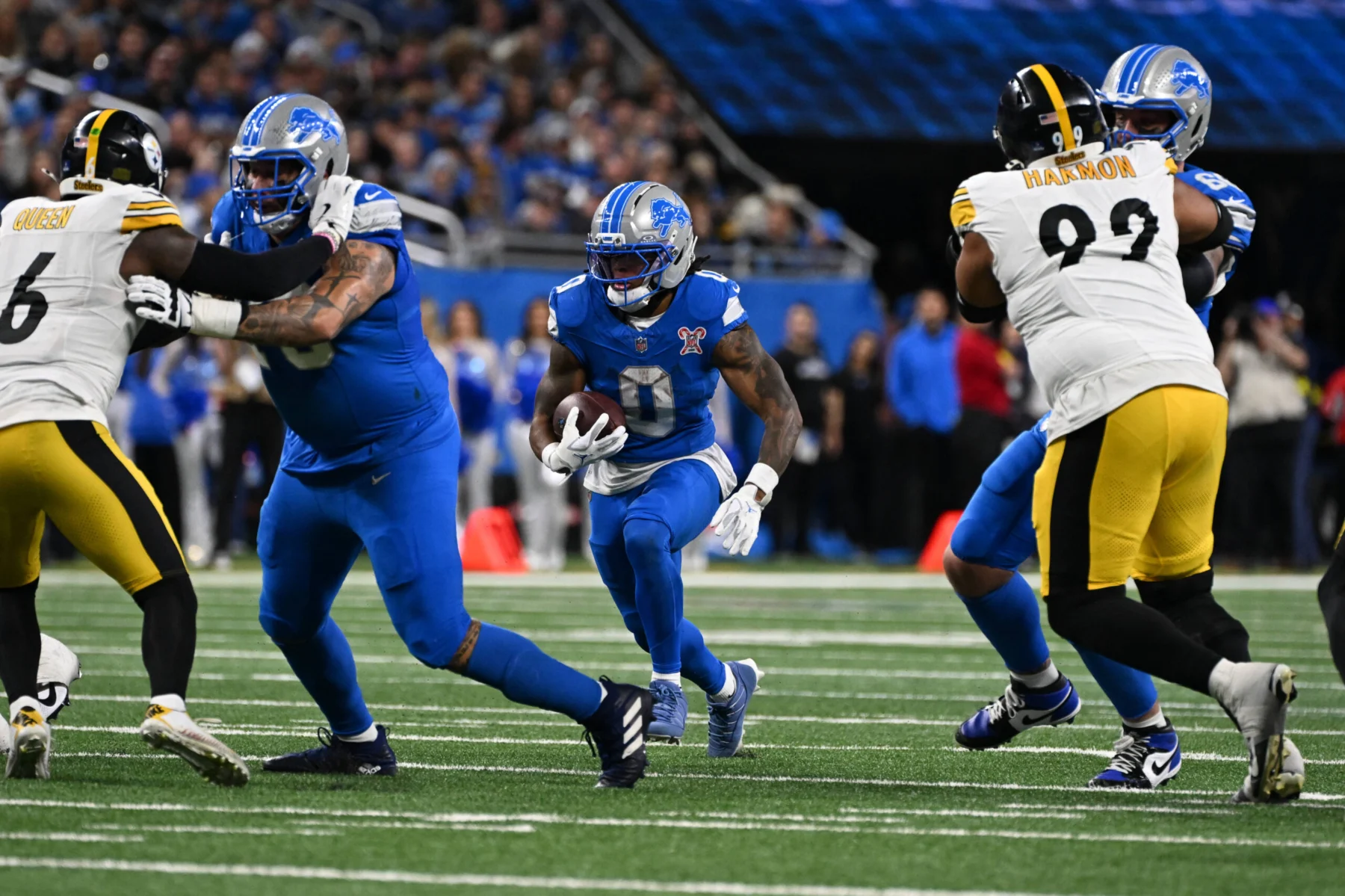 Jahmyr Gibbs, ein Runningback der Detroit Lions, läuft mit dem Football, während seine Teamkollegen und die Verteidiger der Pittsburgh Steelers während eines NFL-Spiels in einen Zweikampf verwickelt sind. Alt-Text wurde KI-generiert.