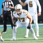 Ein Footballspieler in einem weiß-goldenen Trikot mit der Nummer 77 steht während eines Spiels in Spielposition auf dem Platz. Dillon Radunz macht sich bereit zum Angriff, während im Hintergrund andere Spieler und ein Schiedsrichter zu sehen sind. Alt-Text wurde KI-generiert.