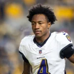 Ein Footballspieler in einem weißen Trikot der Baltimore Ravens mit der Nummer 4 – wahrscheinlich der aufstrebende Star Zay Flowers – steht auf dem Spielfeld, im Hintergrund sind verschwommene Stadionplätze zu sehen. Alt-Text wurde KI-generiert.