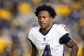 Ein Footballspieler in einem weißen Trikot der Baltimore Ravens mit der Nummer 4 – wahrscheinlich der aufstrebende Star Zay Flowers – steht auf dem Spielfeld, im Hintergrund sind verschwommene Stadionplätze zu sehen. Alt-Text wurde KI-generiert.