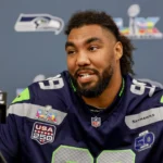 Ein Footballspieler im Trikot der Seattle Seahawks spricht auf einer Pressekonferenz, im Hintergrund sind die Logos der NFL und des Super Bowl zu sehen. Der Bericht „Seahawks verlängern mit Williams“ deutet darauf hin, dass das Team entschlossen ist, seinen Kader zu verstärken. Alt-Text wurde KI-generiert.