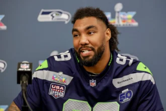 Ein Footballspieler im Trikot der Seattle Seahawks spricht auf einer Pressekonferenz, im Hintergrund sind die Logos der NFL und des Super Bowl zu sehen. Der Bericht „Seahawks verlängern mit Williams“ deutet darauf hin, dass das Team entschlossen ist, seinen Kader zu verstärken. Alt-Text wurde KI-generiert.
