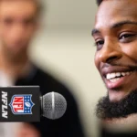 Ein bärtiger Mann lächelt, während er bei einer Presseveranstaltung der Seahawks in ein Mikrofon mit NFL-Logo spricht – möglicherweise über Witherspoons Vertrag –, im Hintergrund ist eine weitere Person unscharf zu sehen. Alt-Text wurde KI-generiert.