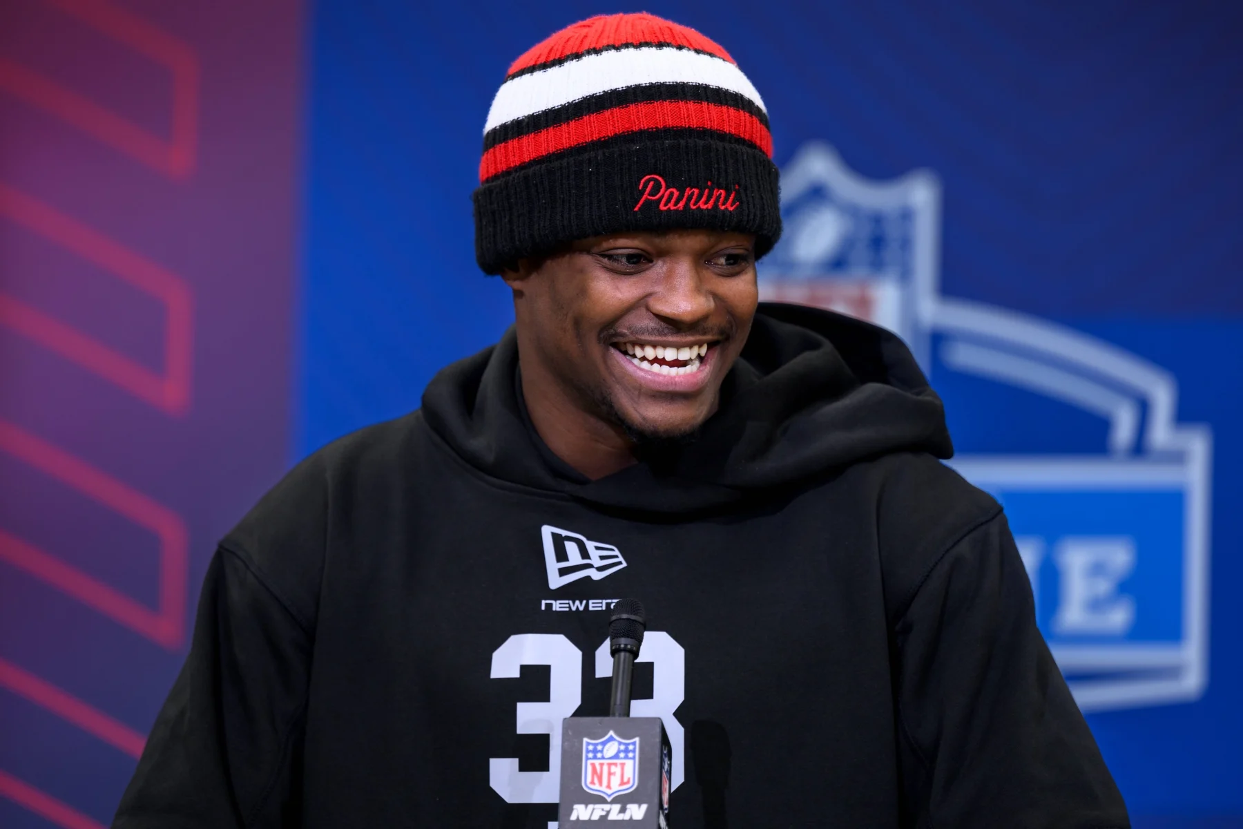 Ein lächelnder Mann, Bud Clark, in einem schwarzen Kapuzenpulli und einer gestreiften Mütze steht auf einem Podium beim NFL Combine, hinter ihm ist ein blau-roter Hintergrund zu sehen. Alt-Text wurde KI-generiert.