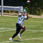 Ein Footballspieler mit weißem Helm und grünem Jets-Trikot mit der Nummer 37, Qwan’tez Stiggers, rennt während des Trainings über das Spielfeld und fängt dabei einen Football. Alt-Text wurde KI-generiert.