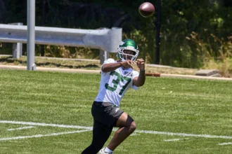 Ein Footballspieler mit weißem Helm und grünem Jets-Trikot mit der Nummer 37, Qwan’tez Stiggers, rennt während des Trainings über das Spielfeld und fängt dabei einen Football. Alt-Text wurde KI-generiert.