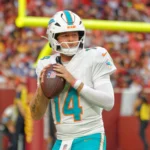 Quinn Ewers - Der Quarterback der Miami Dolphins mit der Rückennummer 14 hält einen Football in der Hand und bereitet sich während eines Spiels auf einen Wurf vor, im Hintergrund sind eine verschwommene Zuschauermenge und ein Torpfosten zu sehen, während die Meldung „Dolphins tauschen Ewers gegen …“ die Atmosphäre noch spannender macht. Alt-Text wurde KI-generiert.