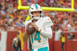 Quinn Ewers - Der Quarterback der Miami Dolphins mit der Rückennummer 14 hält einen Football in der Hand und bereitet sich während eines Spiels auf einen Wurf vor, im Hintergrund sind eine verschwommene Zuschauermenge und ein Torpfosten zu sehen, während die Meldung „Dolphins tauschen Ewers gegen …“ die Atmosphäre noch spannender macht. Alt-Text wurde KI-generiert.