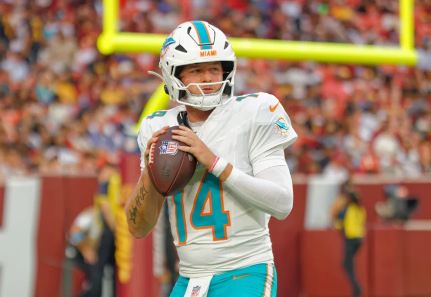Quinn Ewers - Der Quarterback der Miami Dolphins mit der Rückennummer 14 hält einen Football in der Hand und bereitet sich während eines Spiels auf einen Wurf vor, im Hintergrund sind eine verschwommene Zuschauermenge und ein Torpfosten zu sehen, während die Meldung „Dolphins tauschen Ewers gegen …“ die Atmosphäre noch spannender macht. Alt-Text wurde KI-generiert.