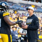 Broderick Jones - Ein Footballspieler in voller Steelers-Uniform reicht einem Mann in Steelers-Fanbekleidung auf dem Spielfeld einen Football, im Hintergrund sind Stadionränge und Zuschauer zu sehen, während ein Bericht meldet, dass die Steelers den ehemaligen Spieler abgeben. Alt-Text wurde KI-generiert.