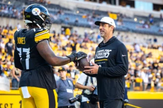Broderick Jones - Ein Footballspieler in voller Steelers-Uniform reicht einem Mann in Steelers-Fanbekleidung auf dem Spielfeld einen Football, im Hintergrund sind Stadionränge und Zuschauer zu sehen, während ein Bericht meldet, dass die Steelers den ehemaligen Spieler abgeben. Alt-Text wurde KI-generiert.