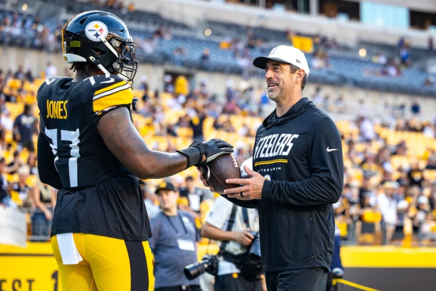 Broderick Jones - Ein Footballspieler in voller Steelers-Uniform reicht einem Mann in Steelers-Fanbekleidung auf dem Spielfeld einen Football, im Hintergrund sind Stadionrรคnge und Zuschauer zu sehen, wรคhrend ein Bericht meldet, dass die Steelers den ehemaligen Spieler abgeben. Alt-Text wurde KI-generiert.