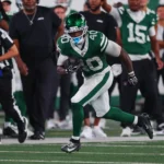 Chip Trayanum - Ein Footballspieler in einem grün-weißen Jets-Trikot läuft während eines Spiels mit dem Ball, während Trainer, Schiedsrichter und Mannschaftskameraden vom Spielfeldrand aus zusehen. Alt-Text wurde KI-generiert.
