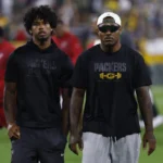 Zwei Männer in Packers-Trikots stehen nebeneinander auf einem Footballfeld, Josh Jacobs trägt eine Sonnenbrille und eine Kappe; im Hintergrund sind verschwommene Menschen und Stadionlichter zu sehen, während LaFleur erwartet, dass die Packers eine starke Saison spielen werden. Alt-Text wurde KI-generiert.