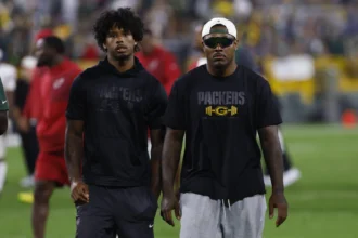 Zwei Männer in Packers-Trikots stehen nebeneinander auf einem Footballfeld, Josh Jacobs trägt eine Sonnenbrille und eine Kappe; im Hintergrund sind verschwommene Menschen und Stadionlichter zu sehen, während LaFleur erwartet, dass die Packers eine starke Saison spielen werden. Alt-Text wurde KI-generiert.
