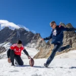 Zwei Männer spielen auf einem verschneiten Berg American Football; einer hält den Ball, während der andere sich darauf vorbereitet, ihn zu kicken, um einen Field-Goal-Weltrekord aufzustellen – vor dem Hintergrund felsiger Gipfel und blauem Himmel –, und dabei die Entschlossenheit von Adam Vinatieri aus seiner legendären Zeit bei den Patriots verkörpern. Alt-Text wurde KI-generiert.