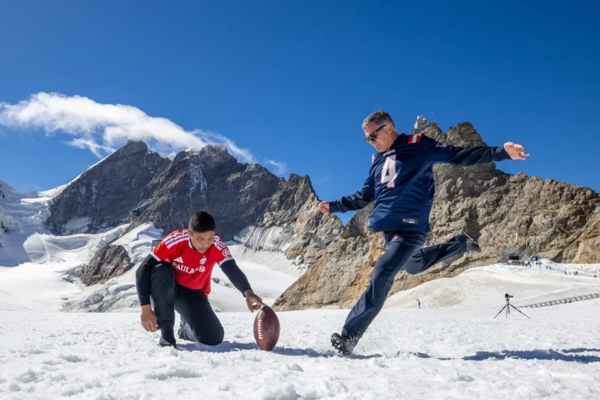 Zwei Männer spielen auf einem verschneiten Berg American Football; einer hält den Ball, während der andere sich darauf vorbereitet, ihn zu kicken, um einen Field-Goal-Weltrekord aufzustellen – vor dem Hintergrund felsiger Gipfel und blauem Himmel –, und dabei die Entschlossenheit von Adam Vinatieri aus seiner legendären Zeit bei den Patriots verkörpern. Alt-Text wurde KI-generiert.