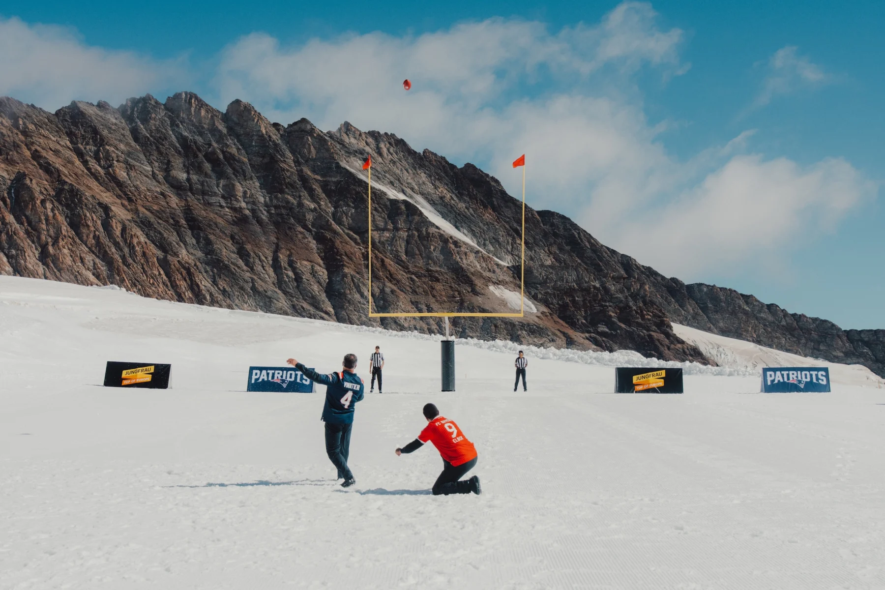 Zwei Menschen spielen Football auf einem verschneiten Feld in der Nähe der Rocky Mountains, im Hintergrund sind zwei Schiedsrichter und „Patriots“-Schilder zu sehen, unter einem teilweise bewölkten blauen Himmel – ein Anblick, der Erinnerungen an Adam Vinatieris legendäre Momente bei Winterspielen weckt. Alt-Text wurde KI-generiert.