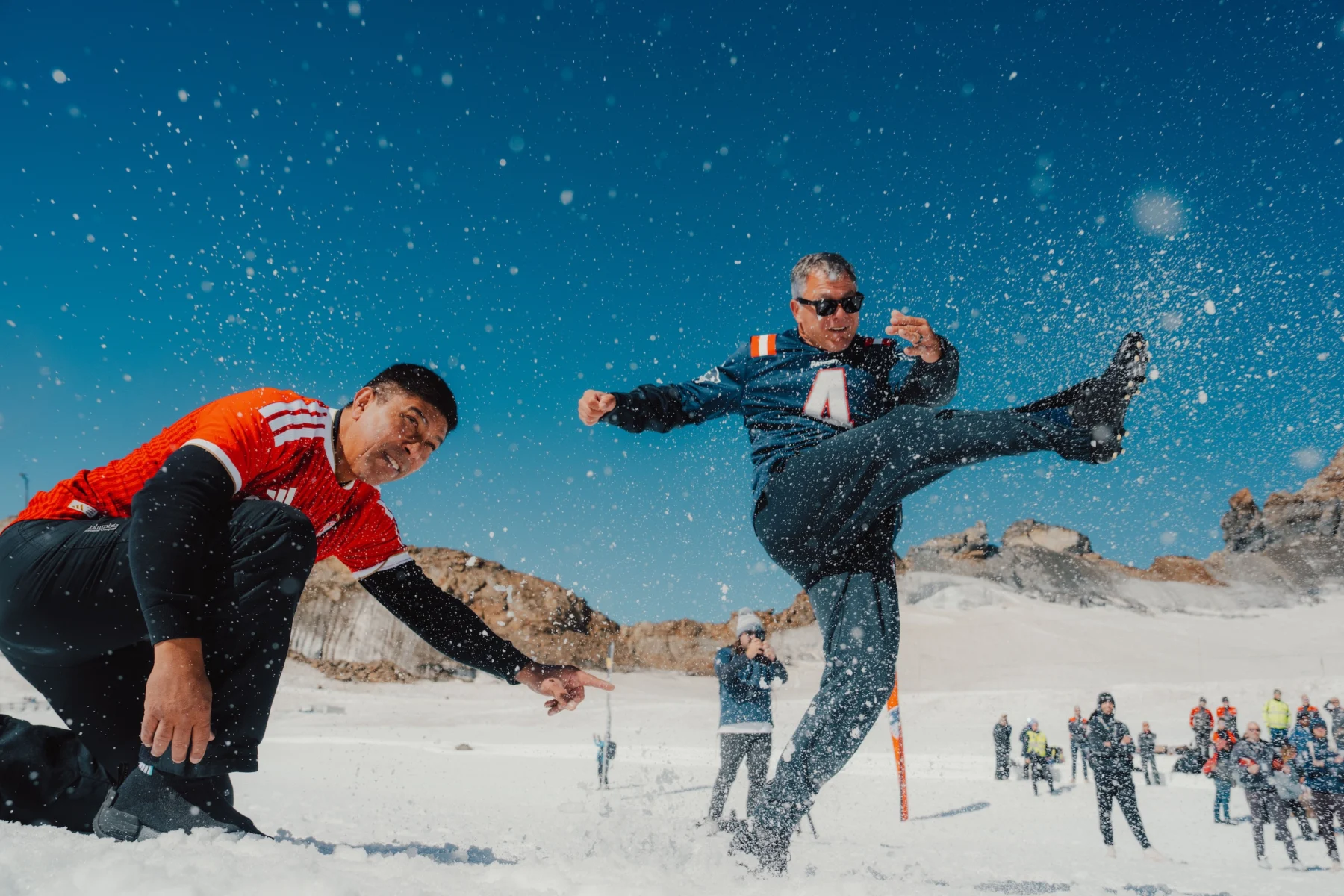Zwei Personen spielen American Football im Schnee: Eine hält den Ball, die andere kickt ihn in einer Pose, die an Adam Vinatieri erinnert. Im verschneiten Hintergrund steht eine Gruppe von Menschen unter einem strahlend blauen Himmel. Alt-Text wurde KI-generiert.