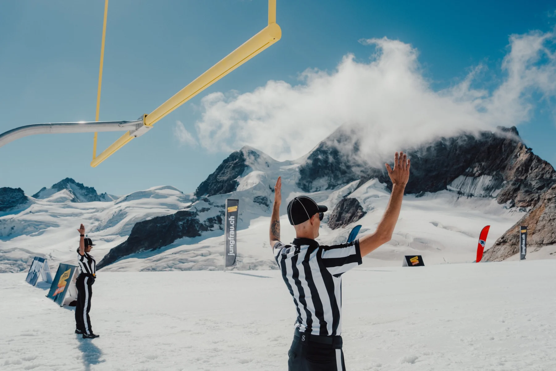 Zwei Fußballschiedsrichter signalisieren ein Field Goal auf einem verschneiten Spielfeld mit Torpfosten, das an Adam Vinatieris legendäre Kicks erinnert, umgeben von Bergen und klarem Himmel. Alt-Text wurde KI-generiert.
