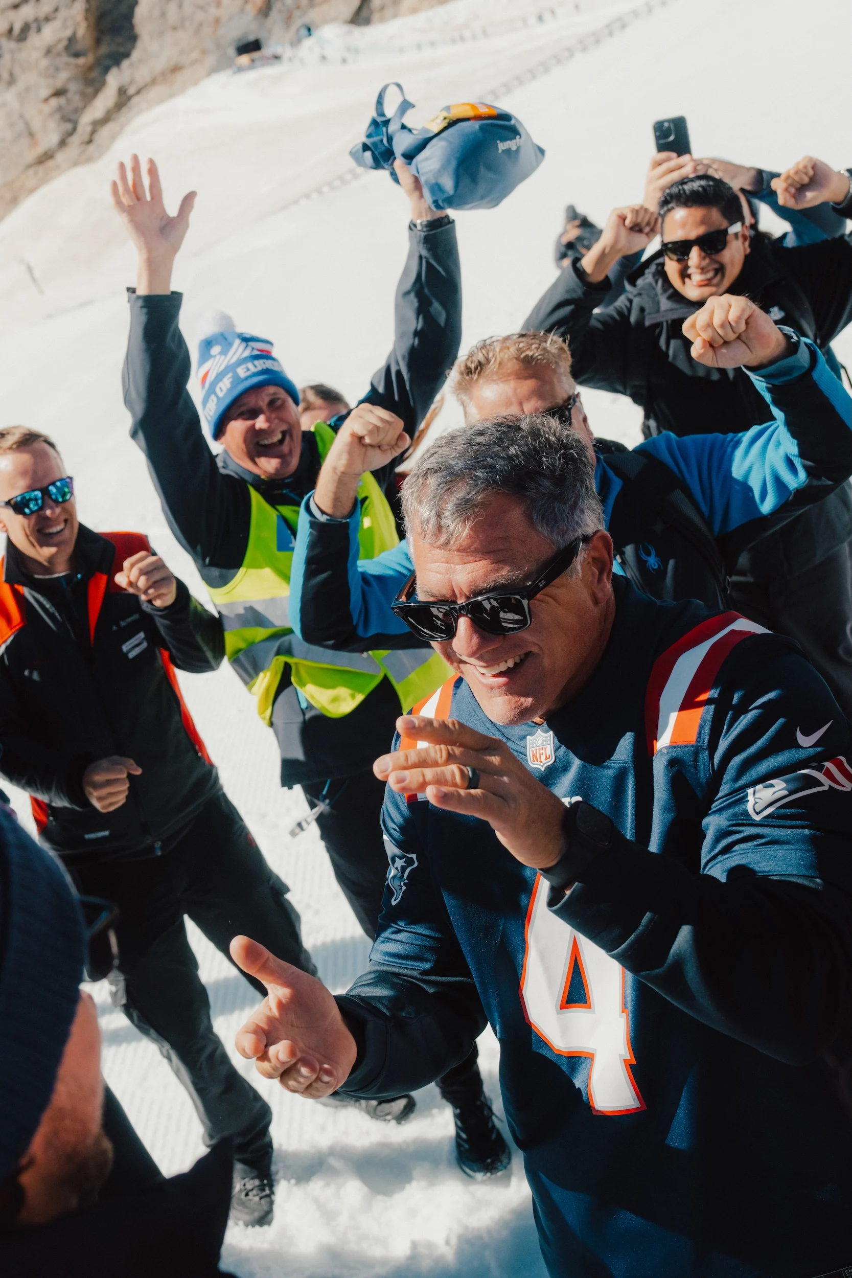 Eine Gruppe von Menschen feiert draußen im Schnee; ganz vorne steht eine Person, die eine Sonnenbrille und ein dunkles Adam-Vinatieri-Football-Trikot trägt. Alt-Text wurde KI-generiert.