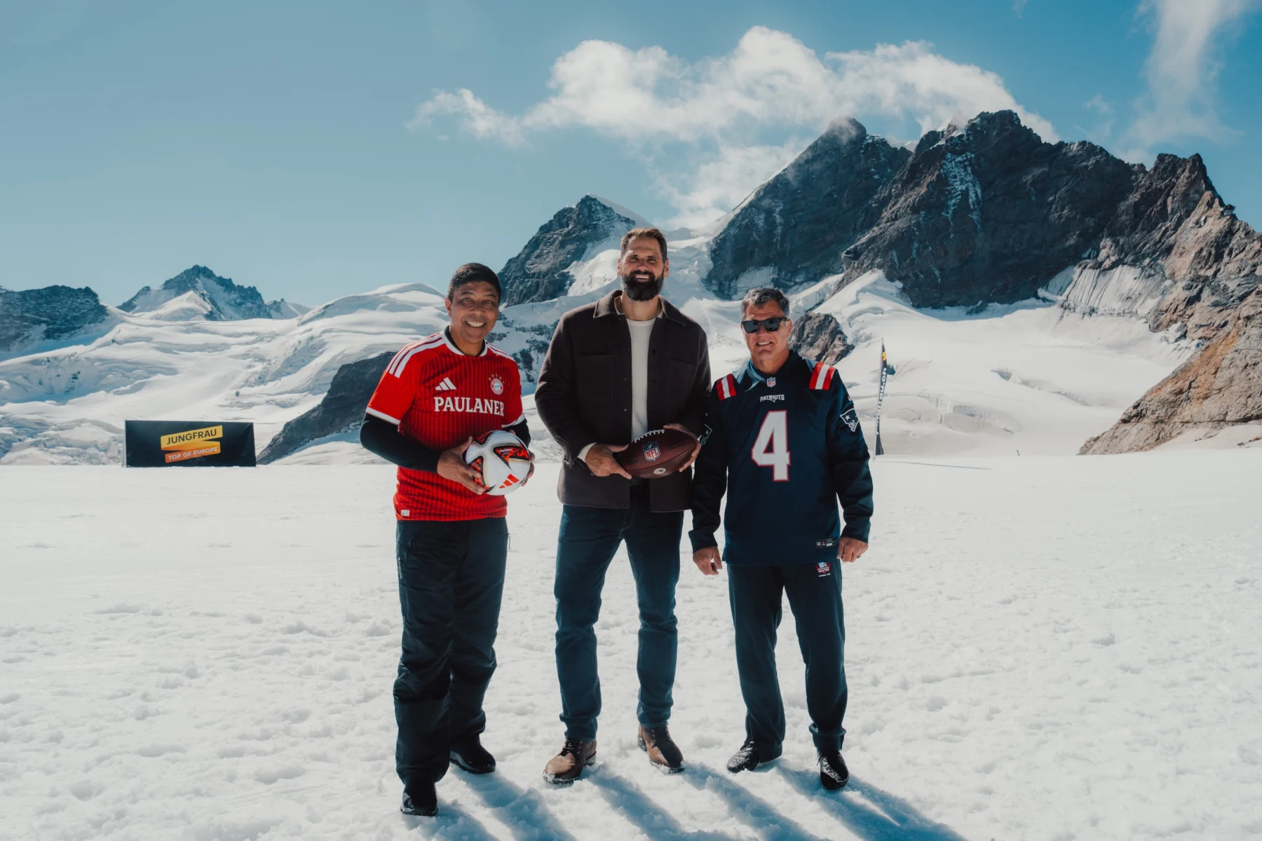 Drei Männer stehen auf einem verschneiten Berg; zwei tragen American-Football-Trikots und halten Footballs in den Händen, während der Mann in der Mitte, Adam Vinatieri, eine Jacke trägt. Im Hintergrund sind schneebedeckte Gipfel und ein gelbes Schild zu sehen. Alt-Text wurde KI-generiert.