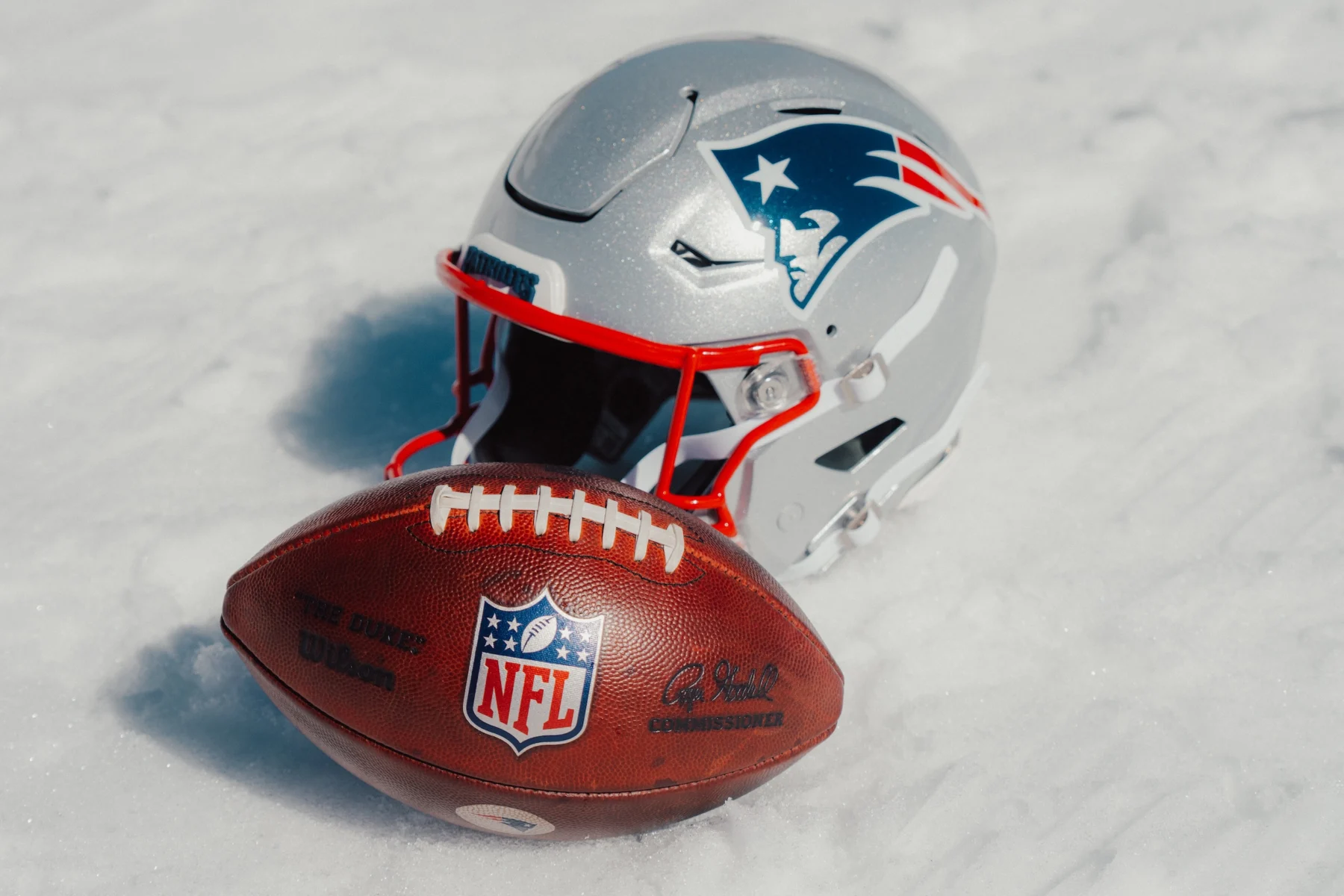 Ein Football-Helm der New England Patriots und ein offizieller NFL-Football liegen im Schnee und wecken Erinnerungen an Adam Vinatieris legendären Kick unter den winterlichen Bedingungen der Playoffs. Alt-Text wurde KI-generiert.