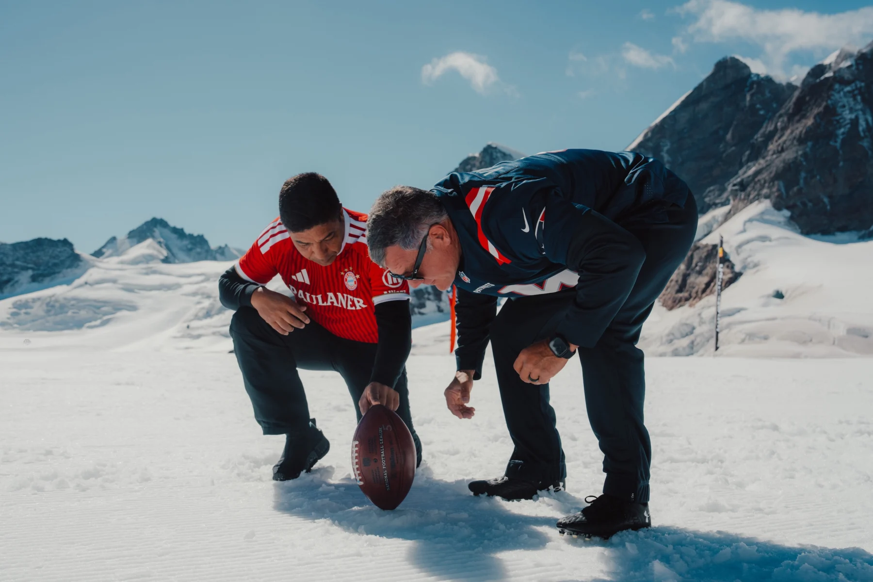 Zwei Männer in Sportkleidung knien im Schnee und legen einen American-Football-Ball auf den Boden, während im Hintergrund schneebedeckte Berge zu sehen sind – eine Nachstellung eines klassischen Field-Goal-Moments von Adam Vinatieri. Alt-Text wurde KI-generiert.