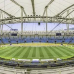 Rio de Janeiro- Weitwinkelaufnahme eines leeren, modernen Stadions mit einem teilweise überdachten Dach, einem grünen Rasenfeld und blauen und gelben Sitzplätzen rund um das Spielfeld; die NFL ist zuversichtlich, dass die Platzverhältnisse für die bevorstehenden Veranstaltungen optimal sind. Alt-Text wurde KI-generiert.