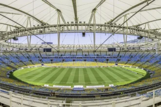 Rio de Janeiro- Weitwinkelaufnahme eines leeren, modernen Stadions mit einem teilweise überdachten Dach, einem grünen Rasenfeld und blauen und gelben Sitzplätzen rund um das Spielfeld; die NFL ist zuversichtlich, dass die Platzverhältnisse für die bevorstehenden Veranstaltungen optimal sind. Alt-Text wurde KI-generiert.