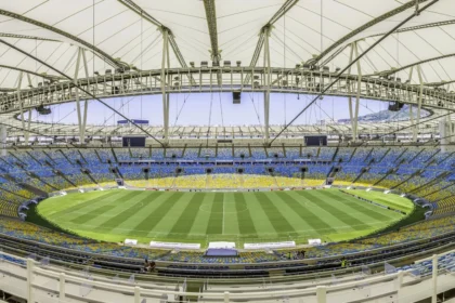 Rio de Janeiro- Weitwinkelaufnahme eines leeren, modernen Stadions mit einem teilweise überdachten Dach, einem grünen Rasenfeld und blauen und gelben Sitzplätzen rund um das Spielfeld; die NFL ist zuversichtlich, dass die Platzverhältnisse für die bevorstehenden Veranstaltungen optimal sind. Alt-Text wurde KI-generiert.