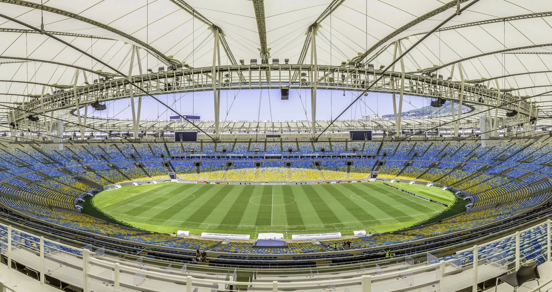 Rio de Janeiro- Weitwinkelaufnahme eines leeren, modernen Stadions mit einem teilweise überdachten Dach, einem grünen Rasenfeld und blauen und gelben Sitzplätzen rund um das Spielfeld; die NFL ist zuversichtlich, dass die Platzverhältnisse für die bevorstehenden Veranstaltungen optimal sind. Alt-Text wurde KI-generiert.