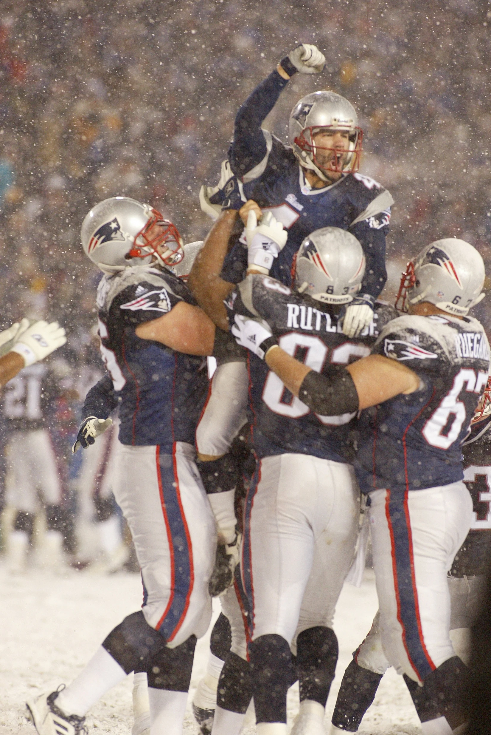 Fußballspieler in den Trikots der New England Patriots heben ihren Teamkollegen Adam Vinatieri während eines Spiels auf einem verschneiten Spielfeld jubelnd in die Höhe. Alt-Text wurde KI-generiert.