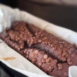 Proteinreiches Schoko-Bananenbrot in Kastenform, garniert mit Schokoladenstückchen, gebacken in einer mit Backpapier ausgelegten Form. Alt-Text wurde KI-generiert.