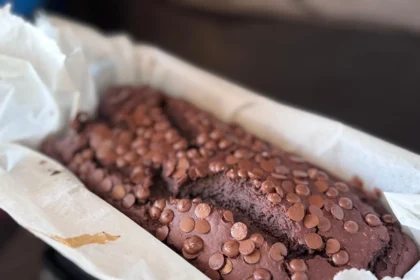 Proteinreiches Schoko-Bananenbrot in Kastenform, garniert mit Schokoladenstückchen, gebacken in einer mit Backpapier ausgelegten Form. Alt-Text wurde KI-generiert.