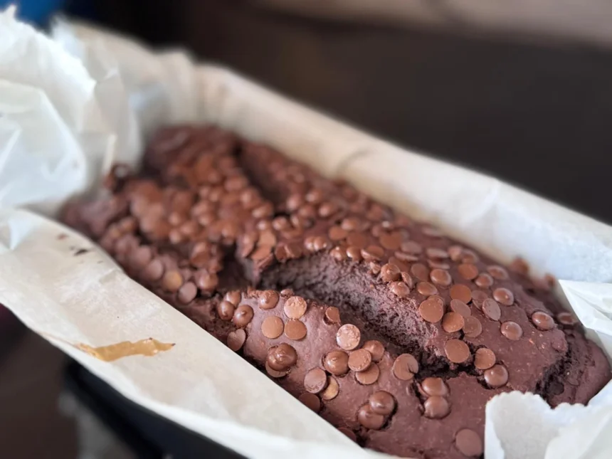 Proteinreiches Schoko-Bananenbrot in Kastenform, garniert mit Schokoladenstückchen, gebacken in einer mit Backpapier ausgelegten Form. Alt-Text wurde KI-generiert.