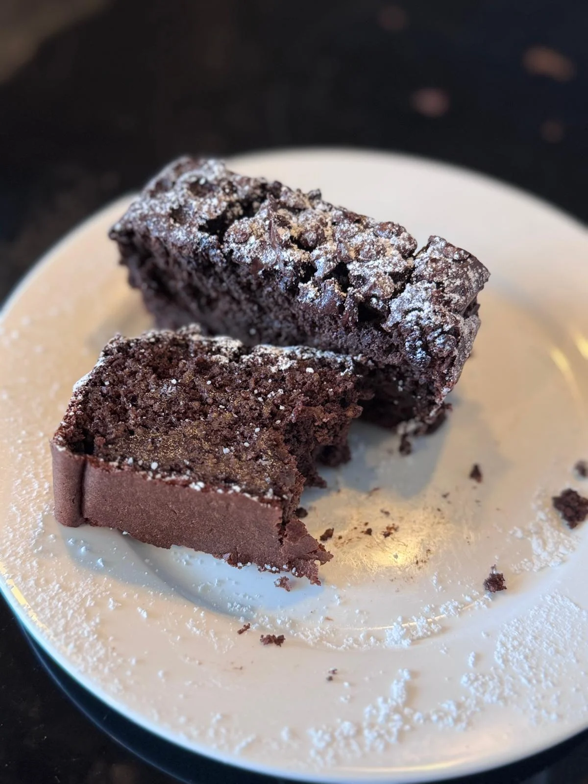 Auf einem weißen Teller liegen zwei Scheiben Schokoladenkuchen mit Puderzucker bestäubt; auf dem Teller sind Krümel zu sehen, die an ein frisch gebackenes proteinreiches Schoko-Bananenbrot erinnern. Alt-Text wurde KI-generiert.