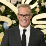 Joe Buck - Ein Mann in einem dunklen Anzug, einem weißen Hemd und einer schwarzen Krawatte steht vor einem blumigen Hintergrund mit goldener Schrift und lächelt in die Kamera – eine Szene, die an eine feierliche Veranstaltung erinnert, bei der Ankündigungen im Stil von „Bericht: Joe Buck einigt sich“ gemacht werden könnten. Alt-Text wurde KI-generiert.