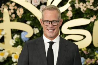 Joe Buck - Ein Mann in einem dunklen Anzug, einem weißen Hemd und einer schwarzen Krawatte steht vor einem blumigen Hintergrund mit goldener Schrift und lächelt in die Kamera – eine Szene, die an eine feierliche Veranstaltung erinnert, bei der Ankündigungen im Stil von „Bericht: Joe Buck einigt sich“ gemacht werden könnten. Alt-Text wurde KI-generiert.