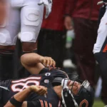 Caleb Williams - Ein Footballspieler im Trikot der Chicago Bears liegt auf dem Spielfeld und hält sich den Arm, während seine Teamkollegen während eines Spiels daneben stehen, nachdem die Bears Williams für verletzt erklärt haben. Alt-Text wurde KI-generiert.